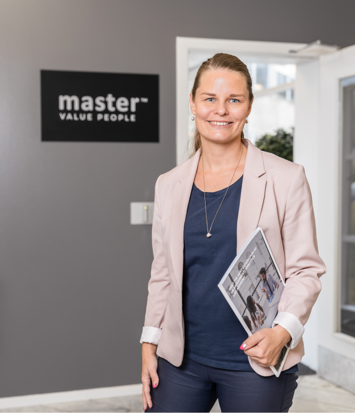 A confident professional woman with blonde hair, wearing a navy blue blouse and a light pink blazer, stands in an office holding a company brochure. Behind her, a sign reads 'master™ VALUE PEOPLE,' reinforcing the organization's mission.