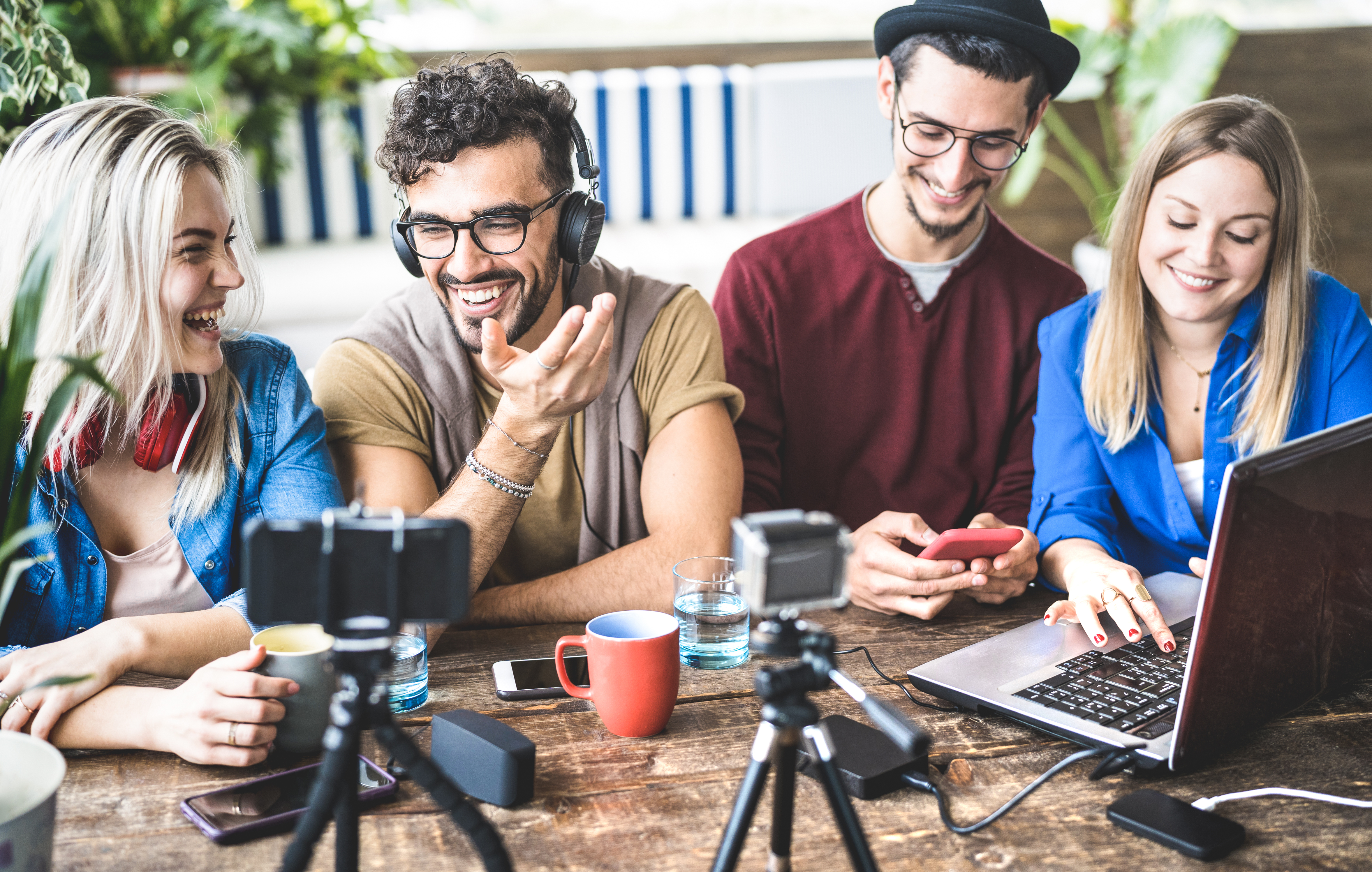  A diverse group of four young adults is gathered around a wooden table, engaged in content creation. They are smiling and laughing while interacting with various digital devices, including a laptop, smartphone, and cameras. A man with curly hair and glasses, wearing headphones, gestures expressively while speaking, while the others look on enthusiastically.