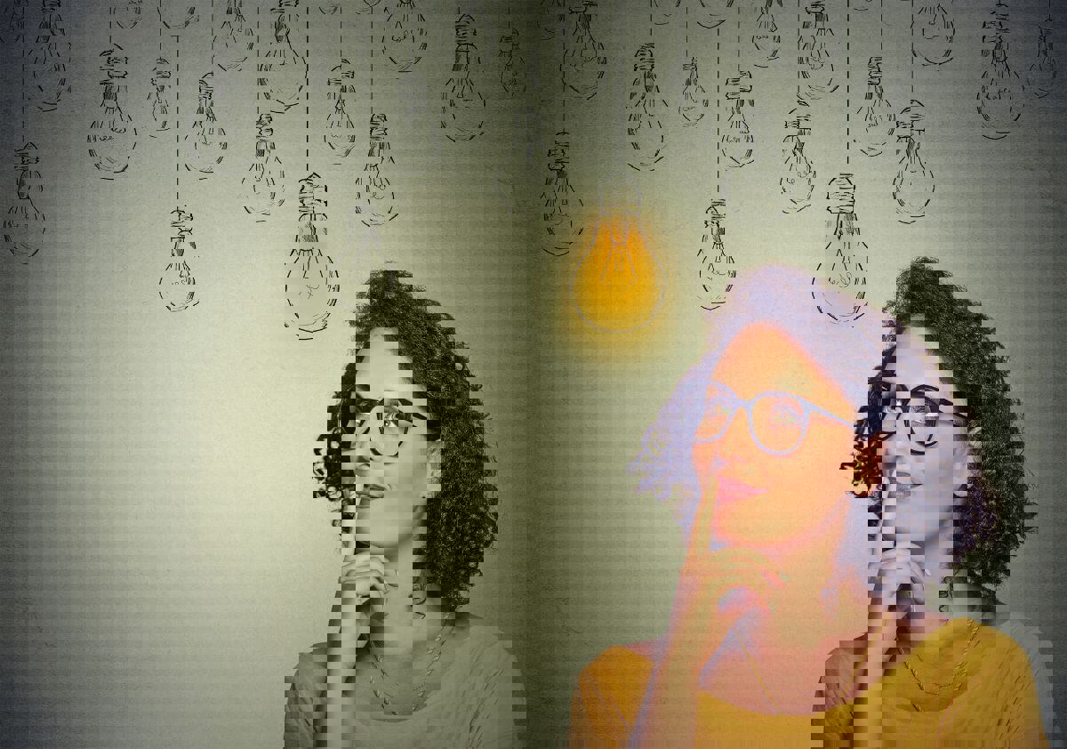  A woman with curly hair and glasses wearing a yellow top is looking up thoughtfully. Above her head, there are multiple hand-drawn lightbulbs hanging, with one glowing yellow, symbolizing an idea or inspiration.