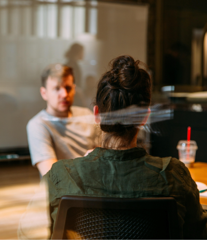 Two professionals are engaged in a focused conversation in a modern office. The woman in the foreground is seen from behind, while the man facing her listens attentively.